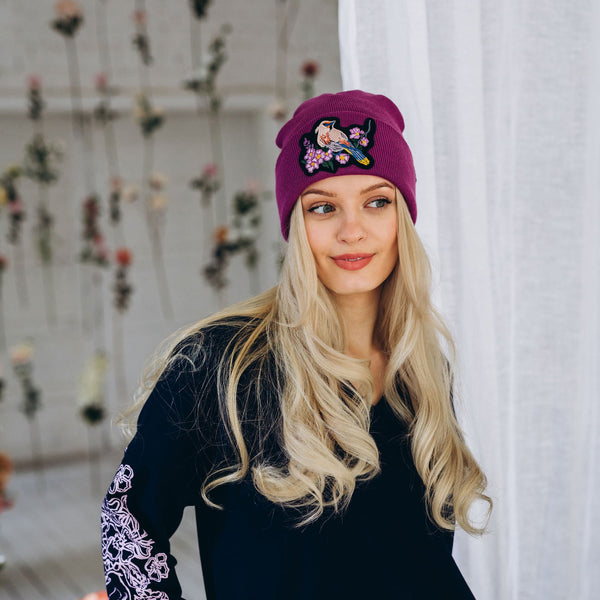 Woman wearing a black dress with floral sleeves and a purple beanie with embroidered bird design